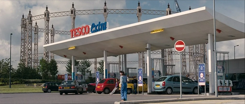 Happy-Go-Lucky (2008) movie still: wide — A Tesco Extra gas station sits beneath a large white canopy near a gasometer. Two people stand near a red…