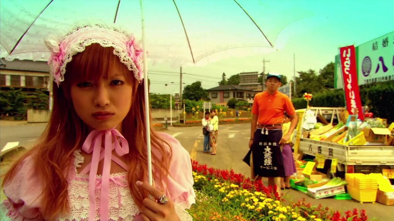 Kamikaze Girls (2004) movie still: closeup — A roadside produce stand sits on a suburban street near a Japanese town. A young woman in a frilly pink…
