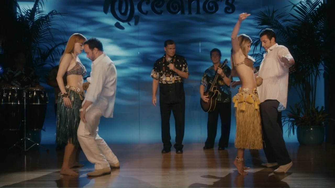 Just Go with It (2011) movie still: fullbody — A hotel stage features two couples performing a hula dance before a blue backdrop. A band and singer…
