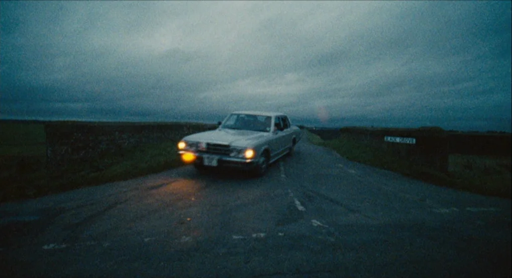 This Is England (2006) movie still: wide — A rural road junction is surrounded by dark grassy embankments under a cloudy sky. A silver vintage car…