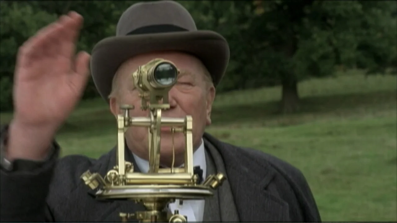 The Gathering Storm (2002) movie still: medium — A man in a grey fedora stands in a grassy field. He looks through a brass surveying theodolite…