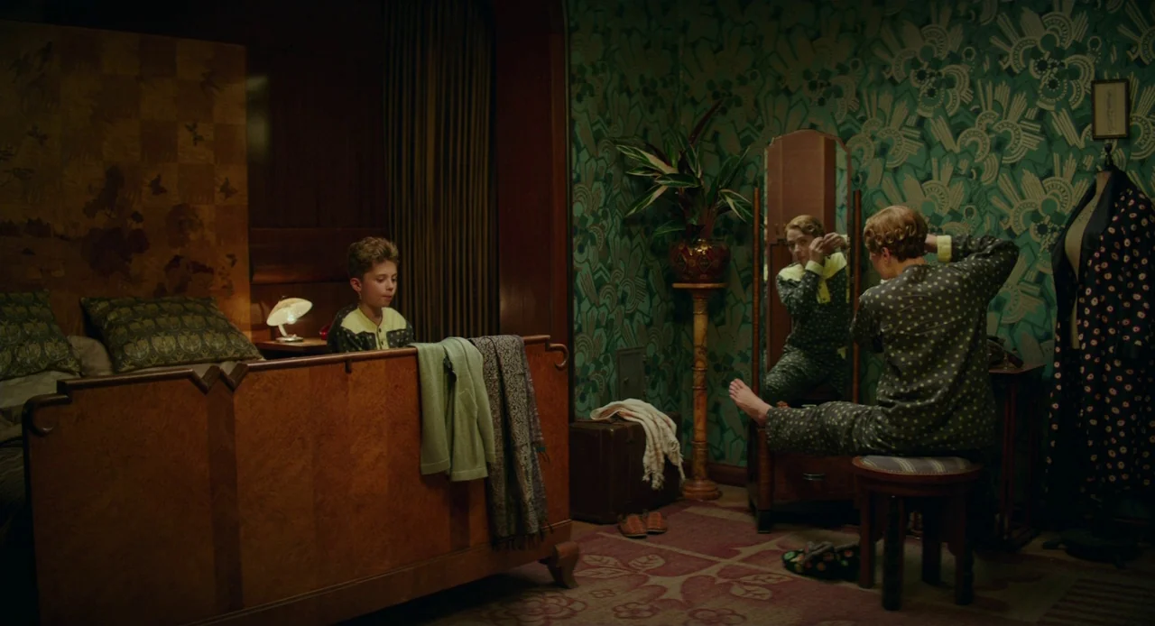 Jojo Rabbit (2019) movie still: wide — A dimly lit room features ornate art deco wallpaper in shades of dark green. A woman prepares for sleep in…