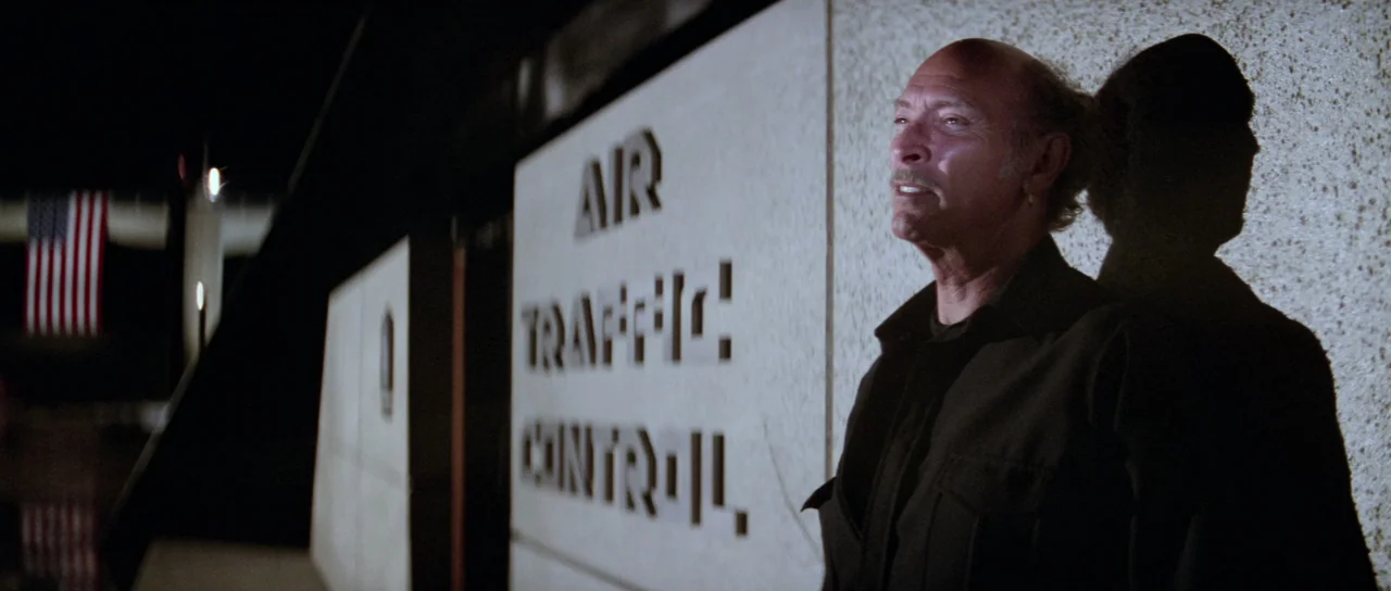 Escape From New York (1981) movie still: medium — An exterior wall at an airport terminal at night featuring prominent signage. A mature man with a…
