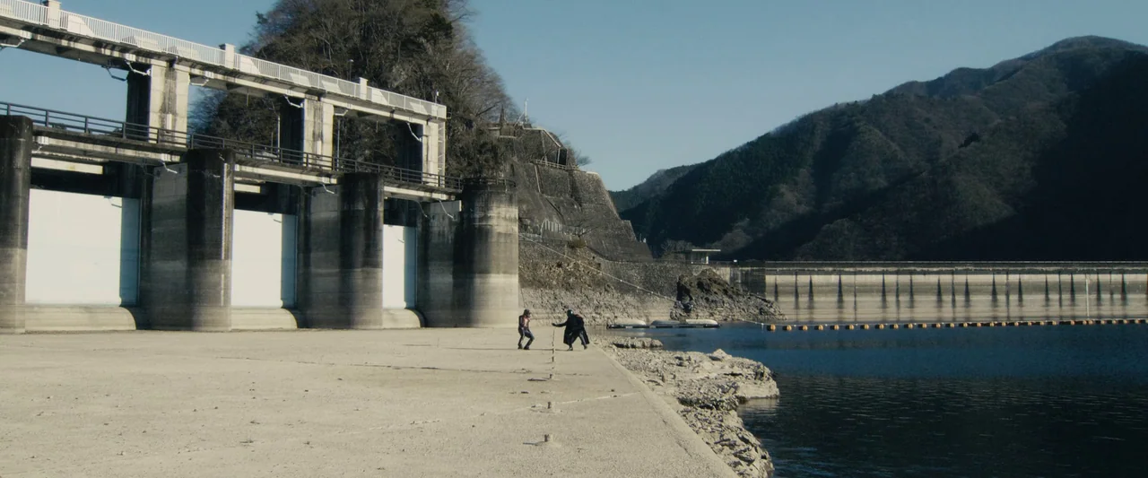 Shin Kamen Rider (2023) movie still: wide — A vast concrete dam structure stands beside a deep blue reservoir with mountain backdrops. Two…