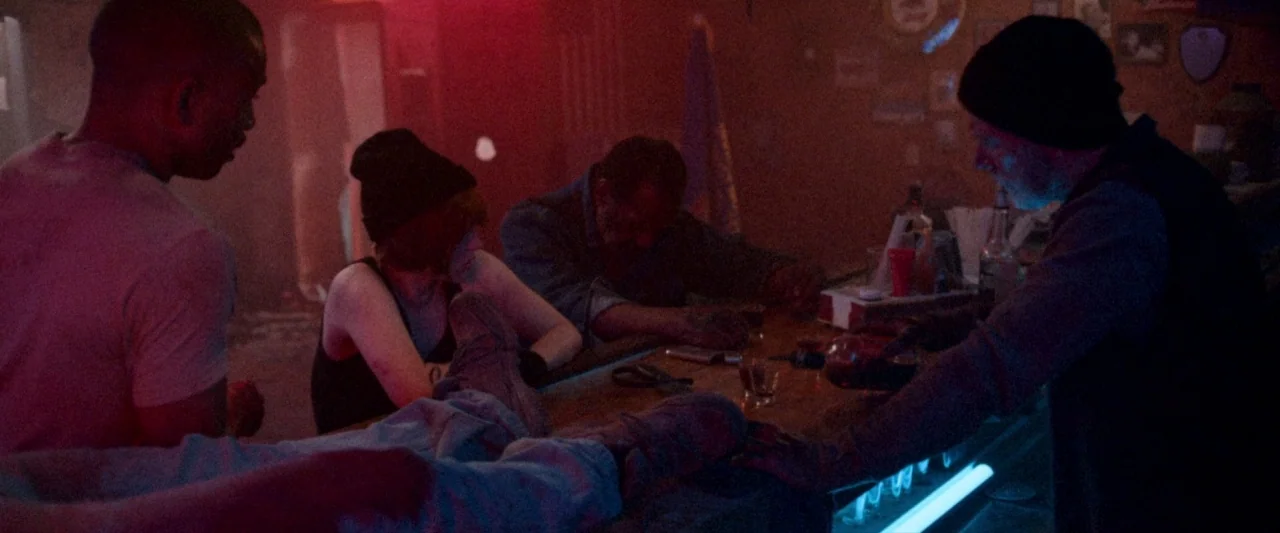 VFW movie still: medium — A dimly lit room with a wooden counter featuring beer signs and liquor bottles. Four people gather around the counter,…