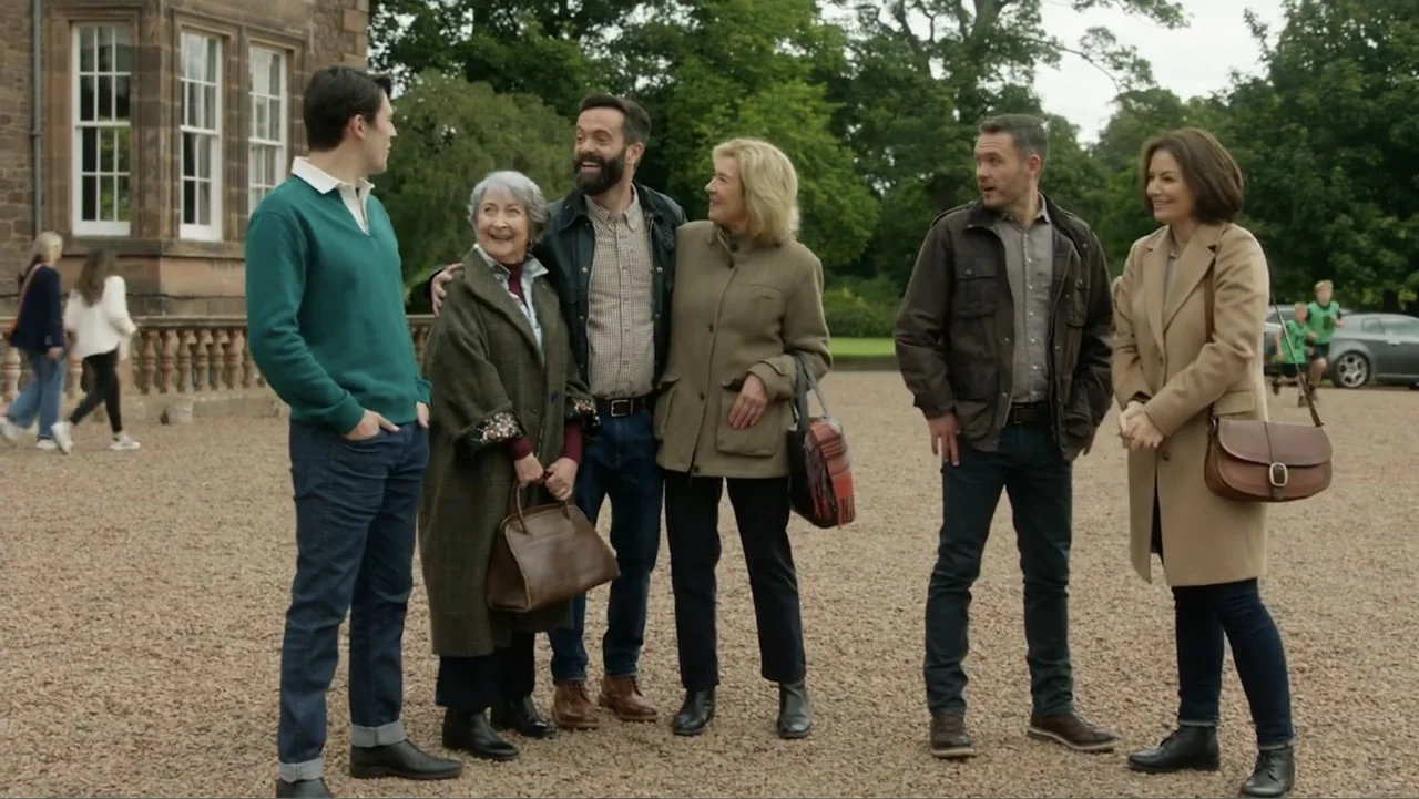 A Scottish Love Scheme (2024) movie still: fullbody — A gravel courtyard sits before a large stone building with tall white-framed windows. Six…