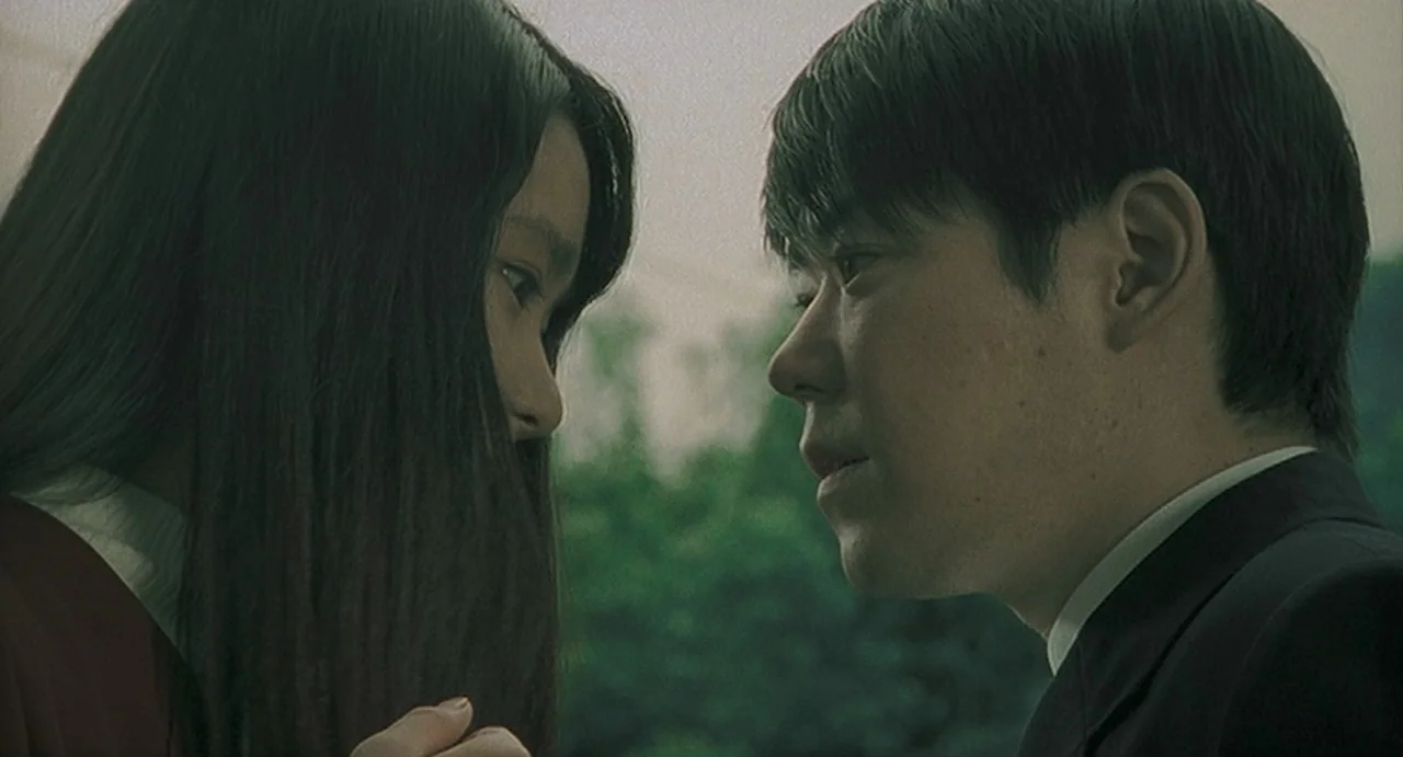 Uzumaki (2000) movie still: closeup — An outdoor area with blurred green foliage visible in the out-of-focus background. Two young adults face each…