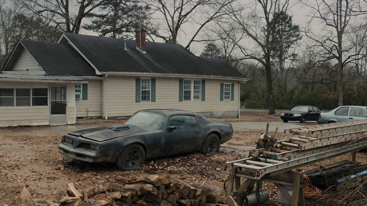 Prisoners (2013) movie still: establishing — A single-story suburban ranch house sits on a dirt lot with bare trees behind. A rusted vintage muscle…