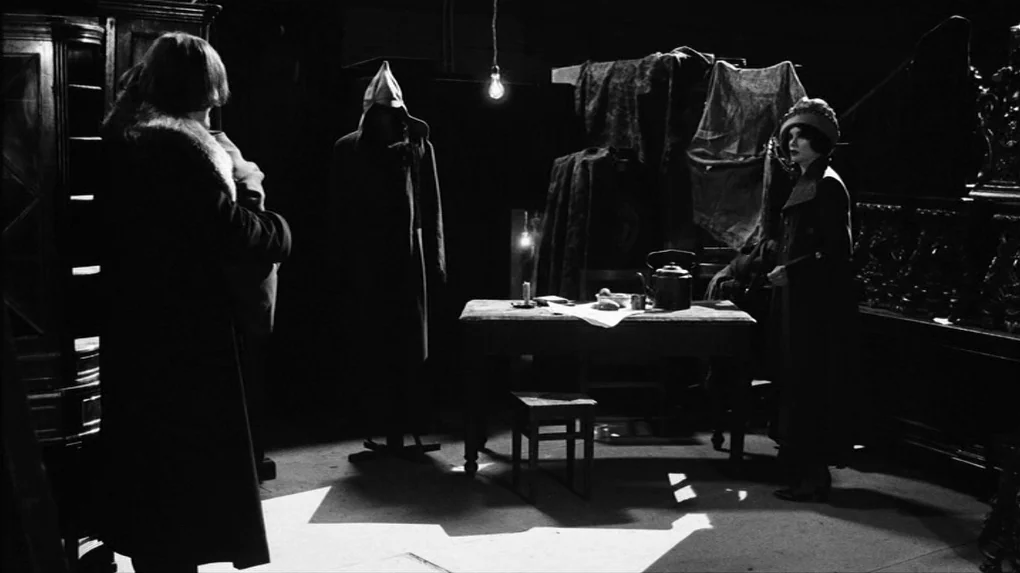 The Role (2013) movie still: wide — A dark sparse room features a central wooden table illuminated by an overhead bulb. Two people stand near the…