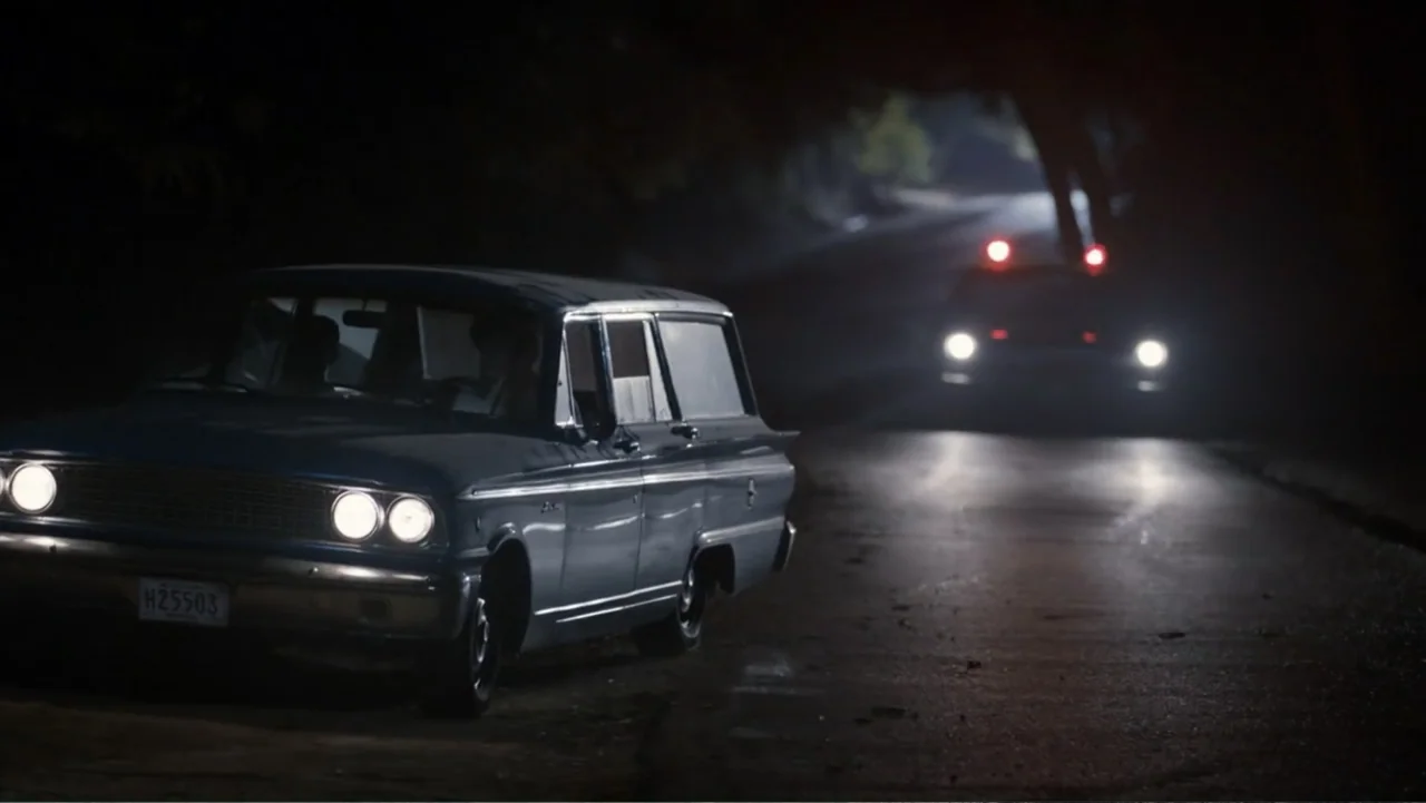 All the Way (2016) movie still: wide — A dark, wooded road is illuminated by the headlights of a vintage station wagon. A second car with visible…