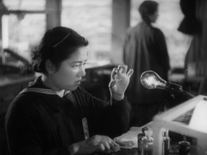 The Most Beautiful (1944) movie still: medium — A woman sits at a wooden table in a dimly lit, cluttered workspace. She examines a small object held…