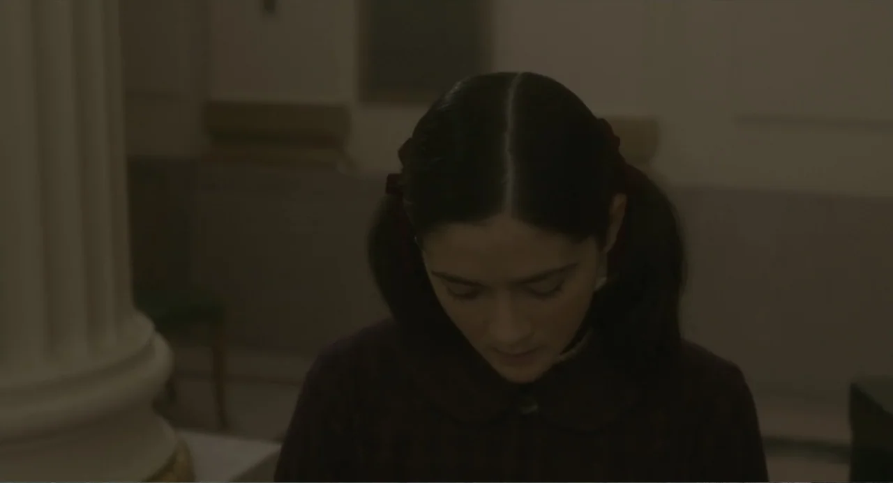 Orphan: First Kill (2022) movie still: closeup — A dimly lit indoor space features a large white architectural column on the left. A young woman…
