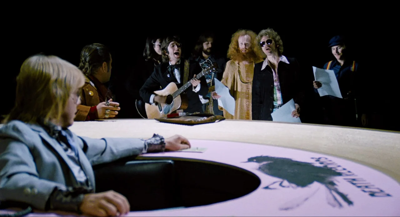 Phantom of the Paradise (1974) movie still: wide — A dimly lit room features a large, light-colored circular desk with a black bird logo. Several…