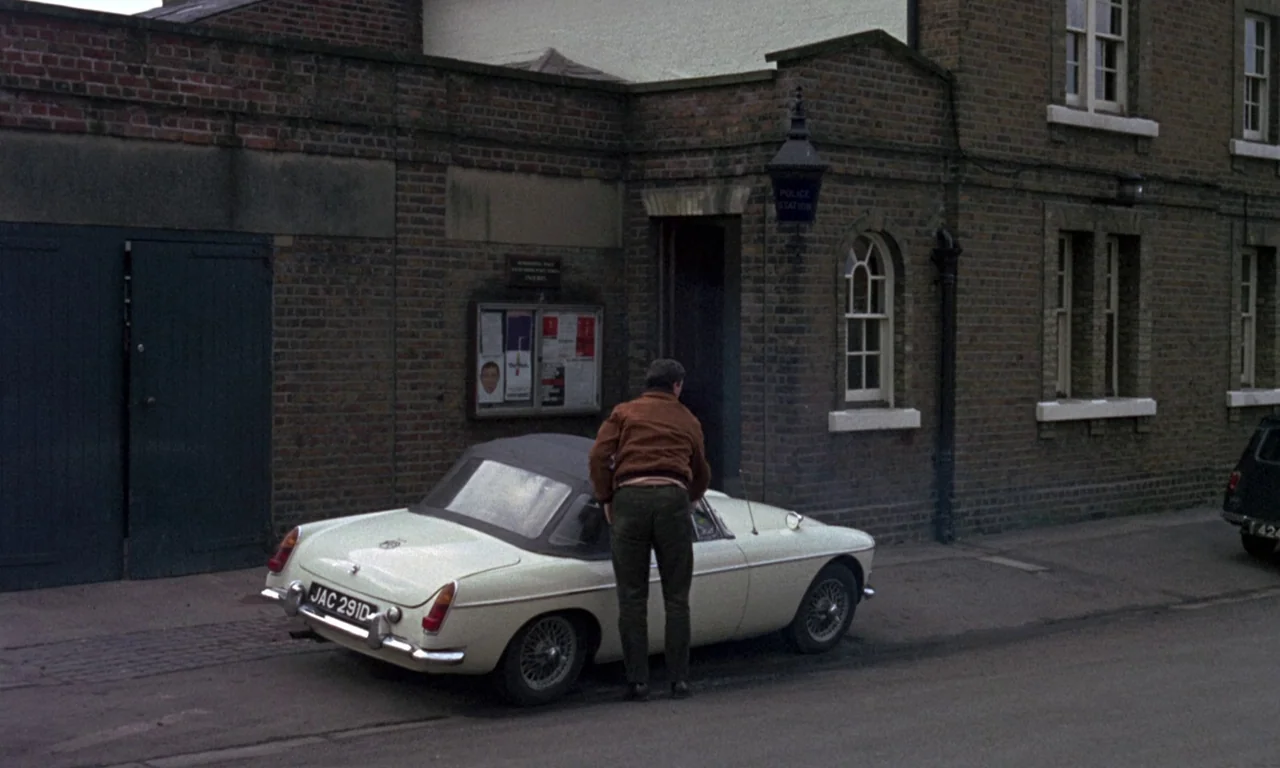 Curse of the Crimson Altar (1968) movie still: fullbody — A brick police station exterior stands beside a paved street with a notice board. A man in…