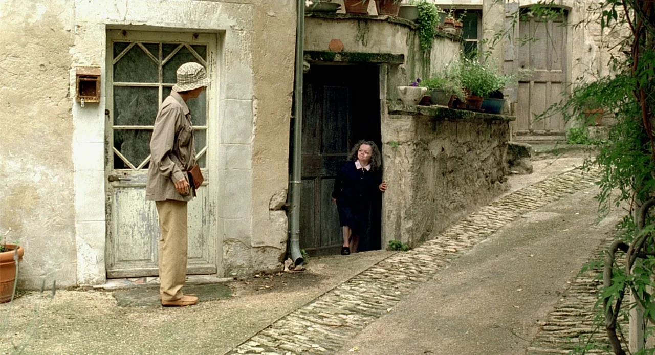 Swimming Pool (2003) movie still: wide — A stone courtyard in a European village features textured walls and a sloped cobblestone path. A man stands…