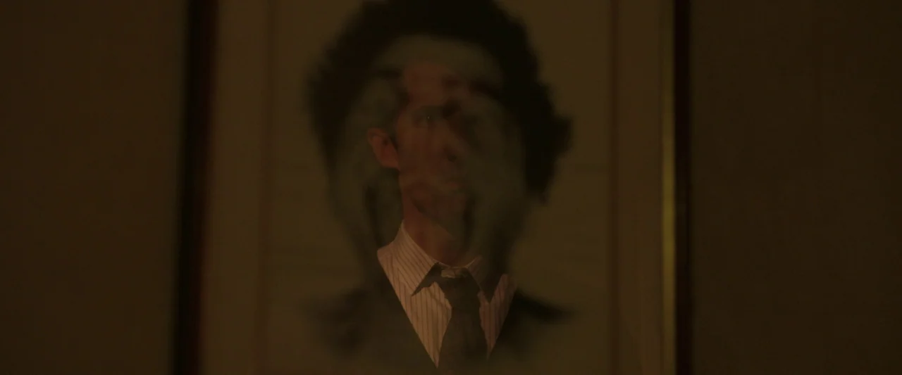 Oddity (2024) movie still: closeup — A man's face appears partially obscured in a dark, reflective surface on a wall. The reflection creates a…