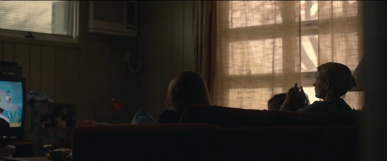 The Good Nurse (2022) movie still: medium — A dimly lit room with a sofa facing a television screen near a window. A woman and a child sit on the…