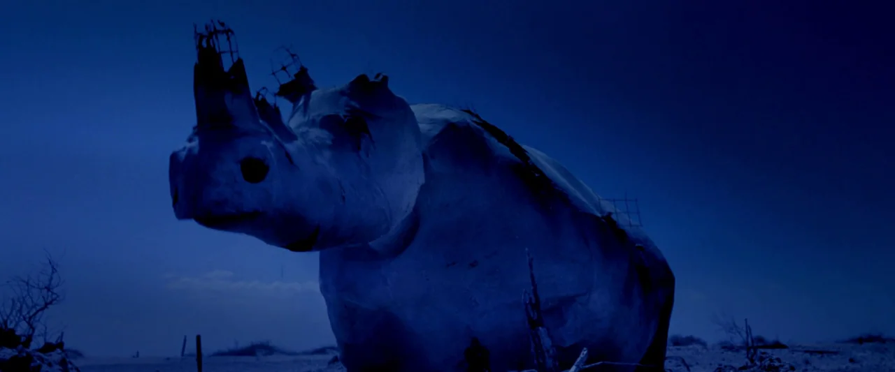 Moon Garden (2022) movie still: medium — A giant, sculptural rhino made of white material stands in a barren desert. The sculpture is partially…