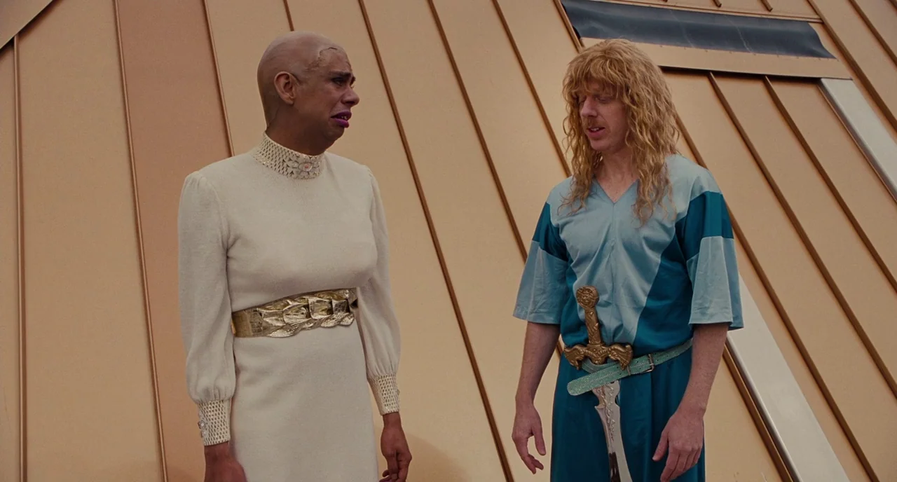 Gentlemen Broncos (2009) movie still: medium — Two individuals stand before a wall clad in vertical gold metallic panels. A bald woman in a white…