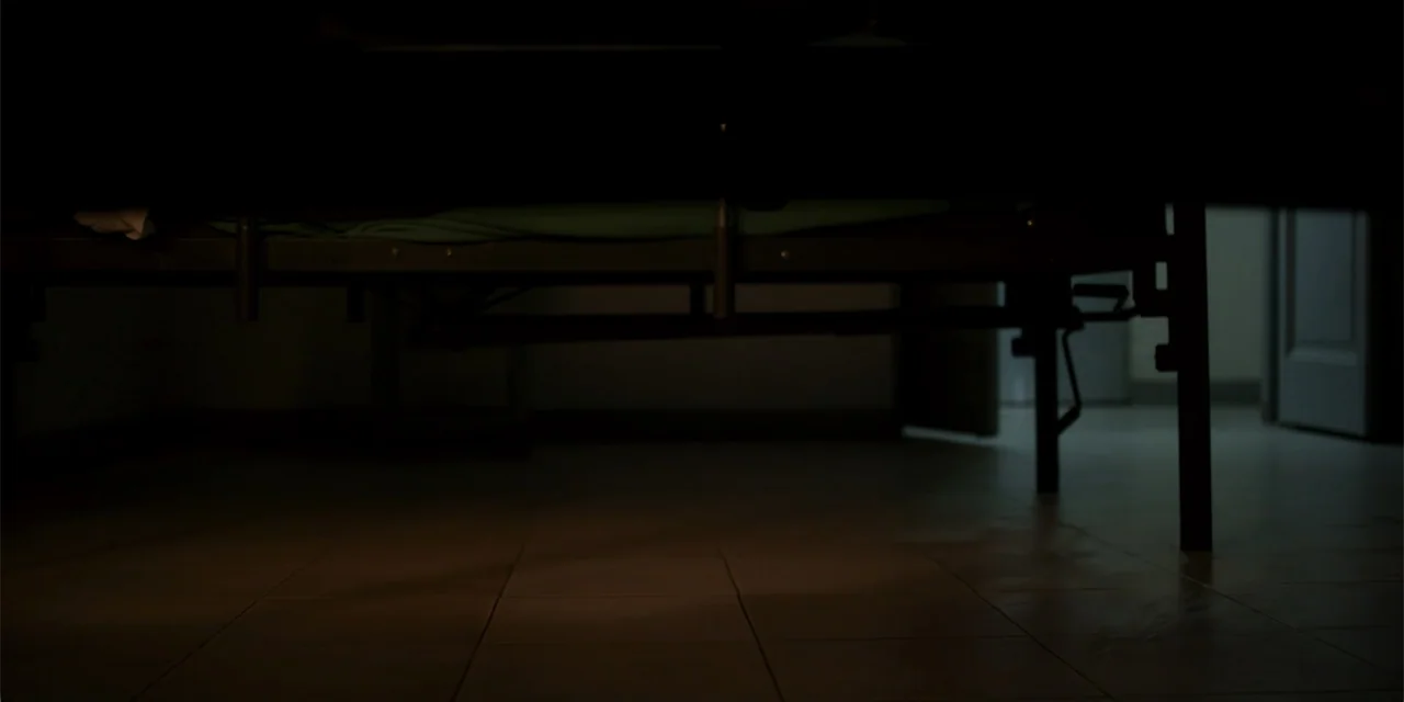 La coda del diavolo (2024) movie still: establishing — A dark hospital room focuses on the space beneath a metal framed bed. The floor is tiled and…