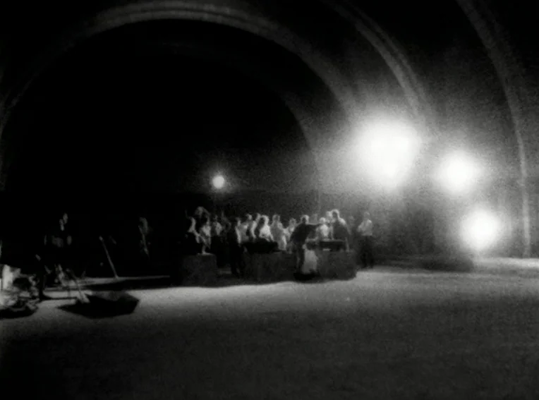 Cuadecuc, Vampir (1971) movie still: wide — A dark, arched-ceiling industrial space with a group standing in the center. Bright, glaring artificial…