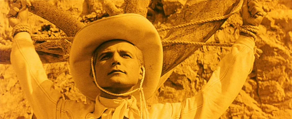 Lemonade Joe (1964) movie still: closeup — A rocky, arid landscape serves as the backdrop for a man bound to wood. The subject faces forward while…