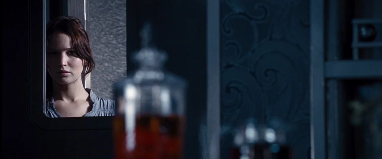 The Hunger Games (2012) movie still: medium — A modern room features dark textured wallpaper and a blurred glass decanter in focus. A young woman…