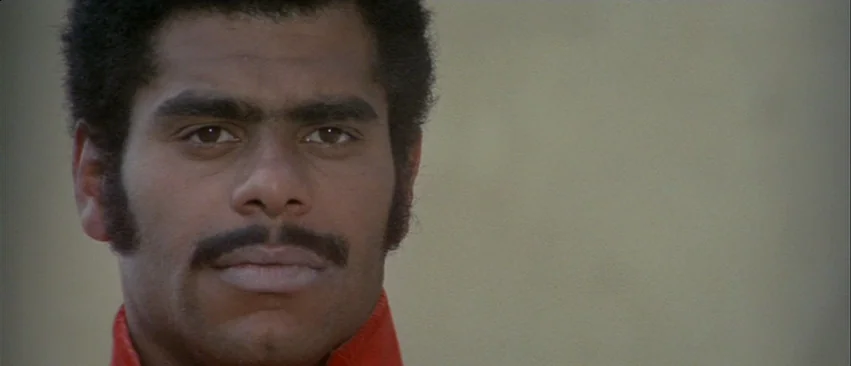 Whity (1971) movie still: closeup — An outdoor area with a smooth, light-colored wall visible in the background. A man with a dark mustache looks…