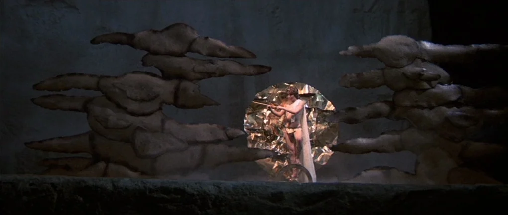 Fellini – Satyricon (1969) movie still: wide — A theatrical set featuring stylized rock-like sculptures flanking a central glowing gold circular…