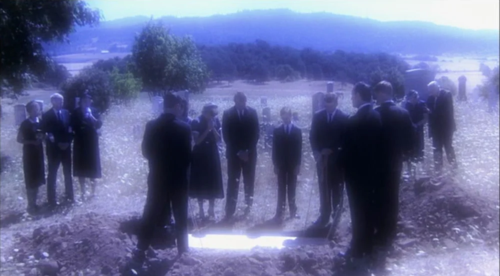 Stand By Me (1986) movie still: wide — A group of people in dark funeral attire stand around an open burial plot. The scene is located in a rural,…