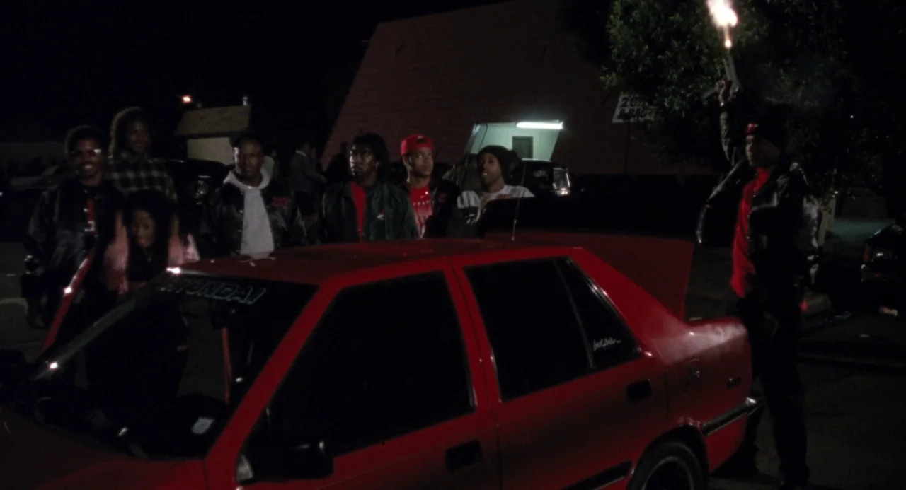 Boyz In The Hood (1991) movie still: wide — A group of young men gathers around a bright red sedan in a dark parking lot at night. One man stands by…