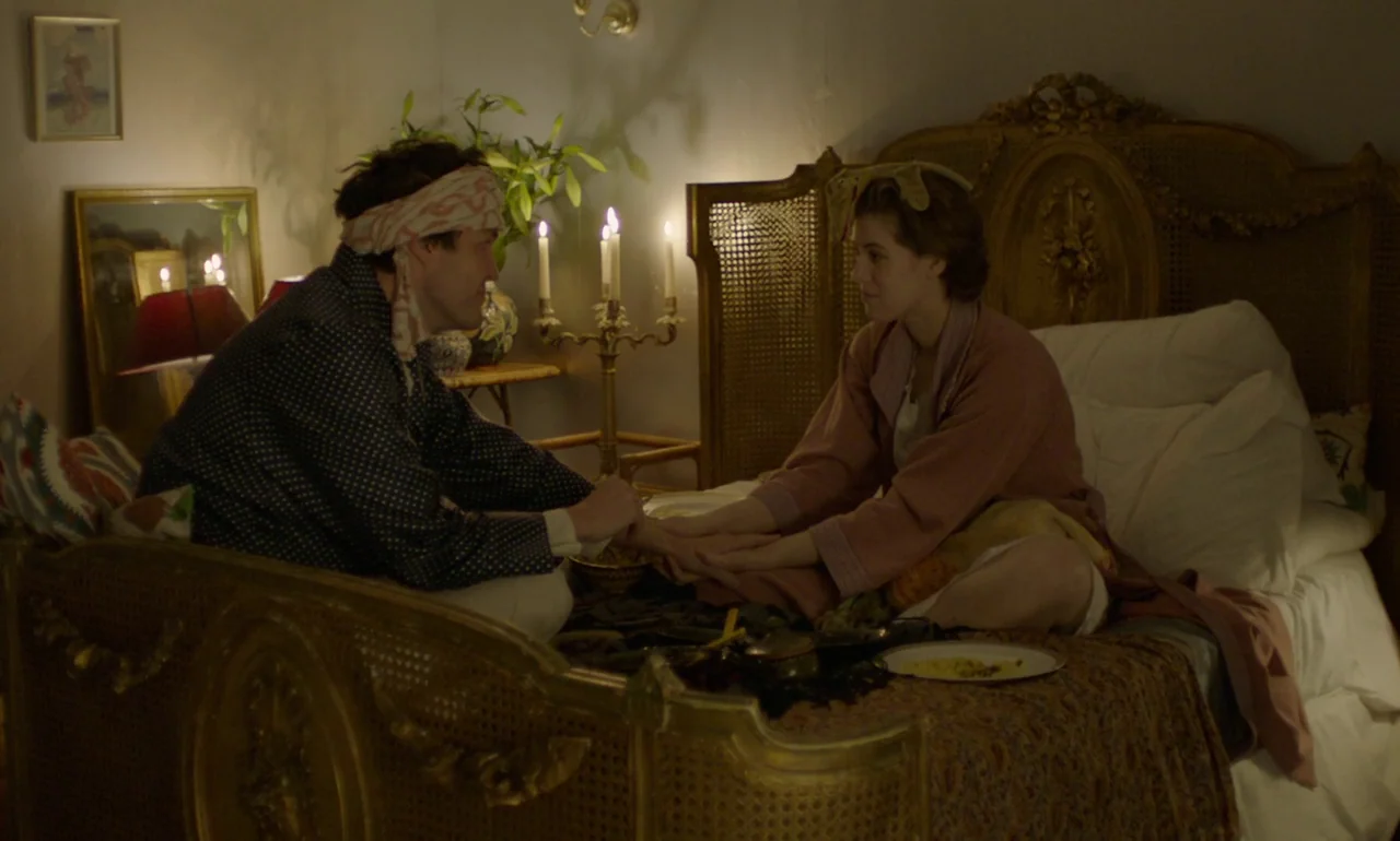 The Souvenir (2019) movie still: medium — A dimly lit room with an ornate carved gold-leaf bed frame and candlelight. A man with a bandaged head and…