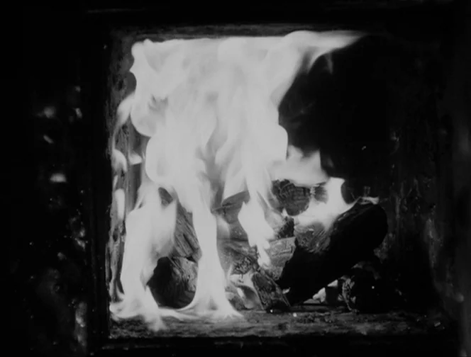 The Ascent (1977) movie still: closeup — A square metal or stone fireplace interior holds burning wood logs. Bright white flames rise upward amidst…
