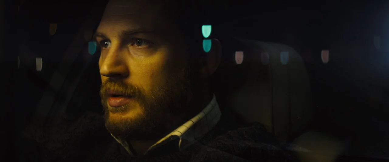 Locke (2013) movie still: closeup — The interior of a car at night with a man sitting behind the wheel. The man looks out the side window as city…