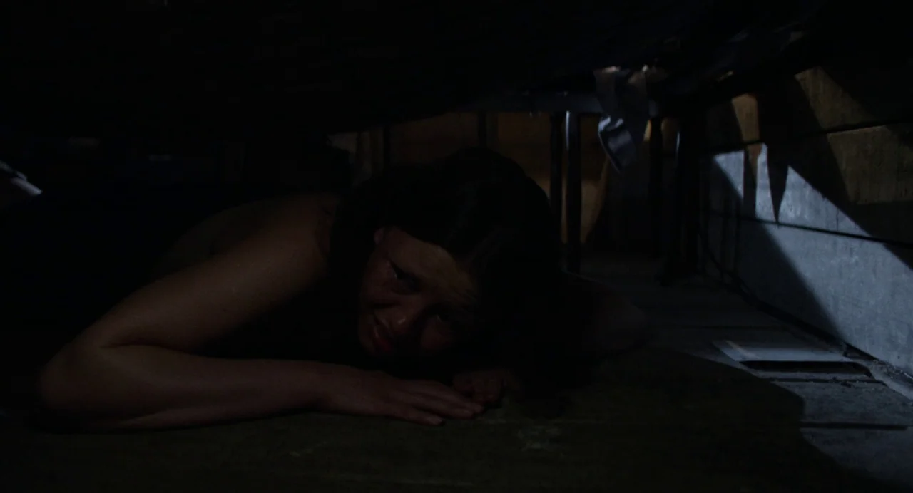 X (2022) movie still: medium — A dark space beneath a metal bed frame with wooden flooring and visible vertical supports. A young woman lies prone…