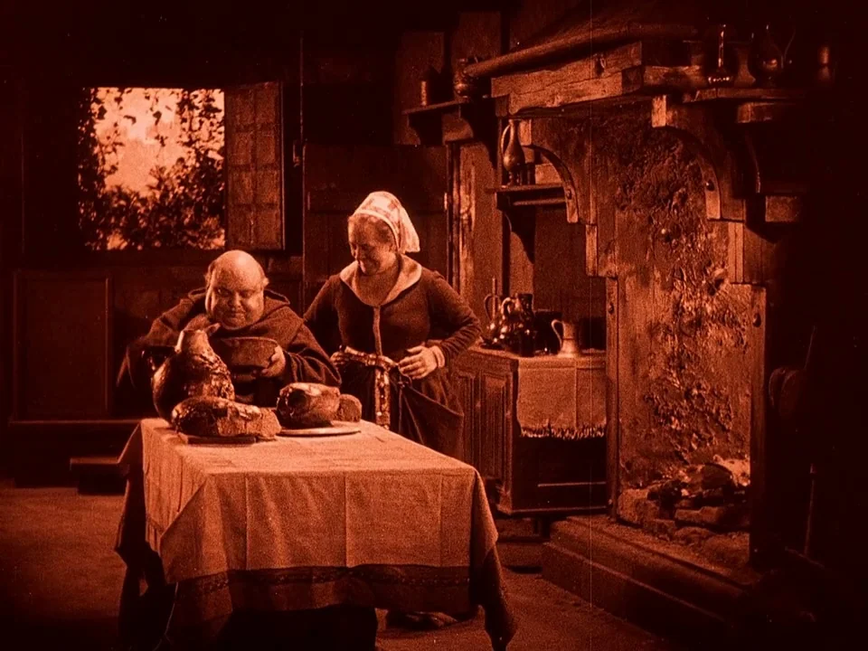 Häxan (1922) movie still: medium — A rustic stone interior features a large fireplace and wooden furniture from a medieval era. A monk consumes a…