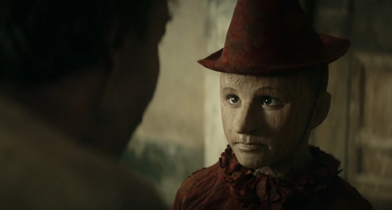 Pinocchio (2022) movie still: medium — A dimly lit interior room features a wooden puppet interacting with a human subject. The puppet wears a red…