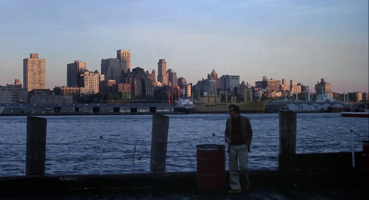 Annie Hall (1977) movie still: wide — A wooden pier extends over a wide river with a cityscape skyline in distance. A man in a jacket stands by a…