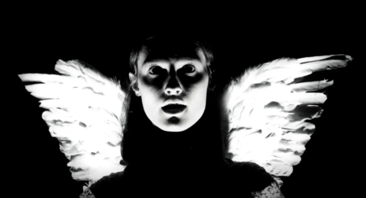 Where The Devil Roams (2023) movie still: closeup — A person stands in a pitch black environment with bright white wings. They face the camera with…