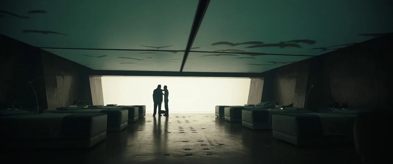 Assassin’s Creed (2016) movie still: wide — A spacious, darkened room with symmetrical rows of minimalist beds and a bright rear window. Two men…