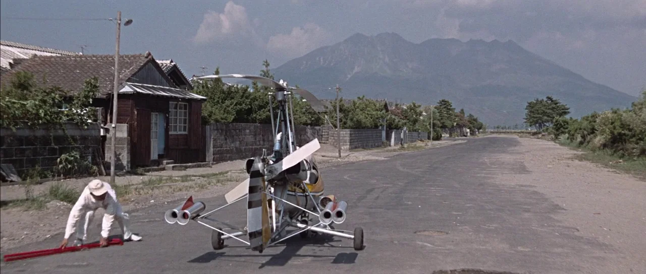 You Only Live Twice (1967) movie still: wide — A rural paved road features a small experimental autogyro vehicle parked in the middle. A man in…