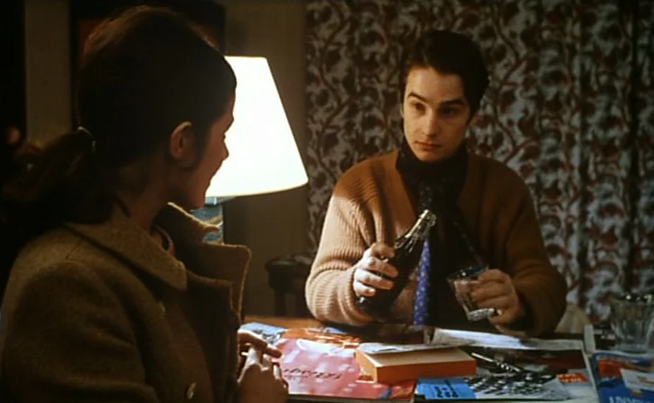 Stolen Kisses (1968) movie still: medium — A dimly lit indoor space featuring a patterned curtain background and a wooden table. A man in a brown…