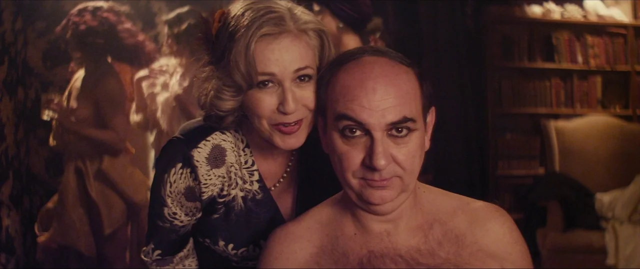 Neruda (2016) movie still: medium — A dimly lit room with a dark wooden bookcase in the back. A blonde woman embraces a shirtless man wearing bold…