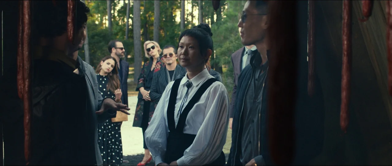 The Menu (2022) movie still: medium — A group of people gathers outdoors in a pine forest setting during the day. They stand together while…