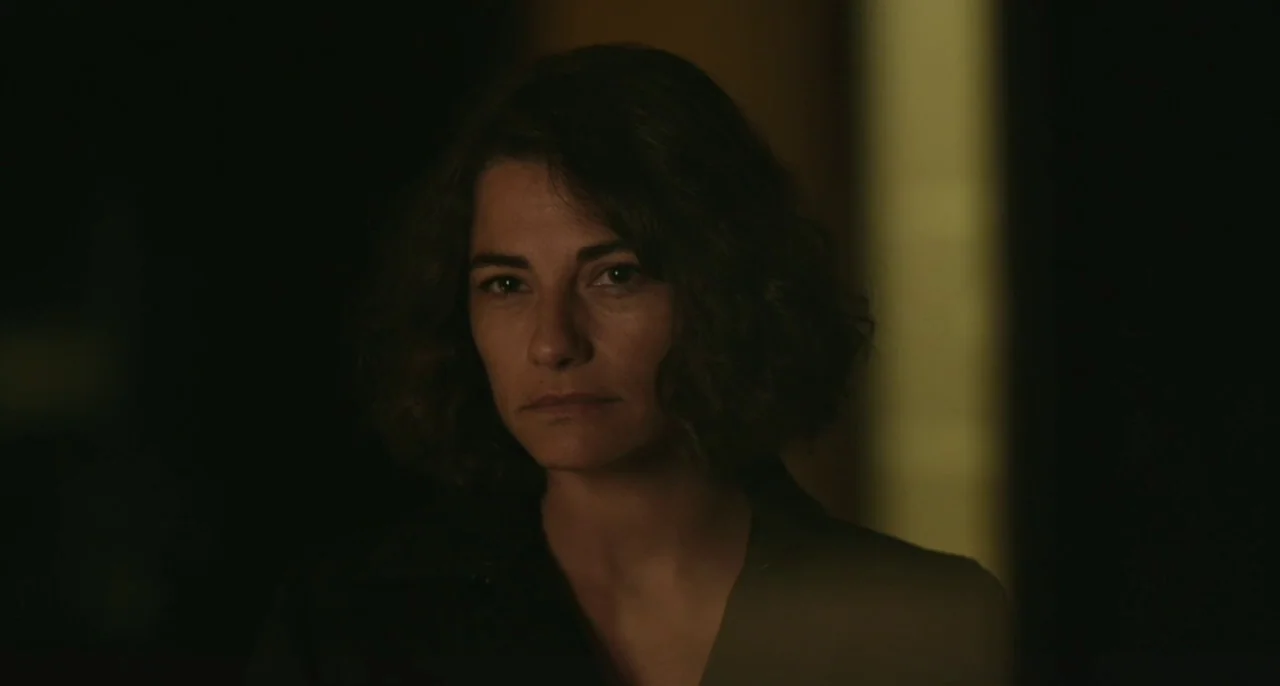 Bastards (2013) movie still: closeup — A woman stands in a dimly lit, dark space with a faint vertical light stripe. She gazes ahead with a neutral…