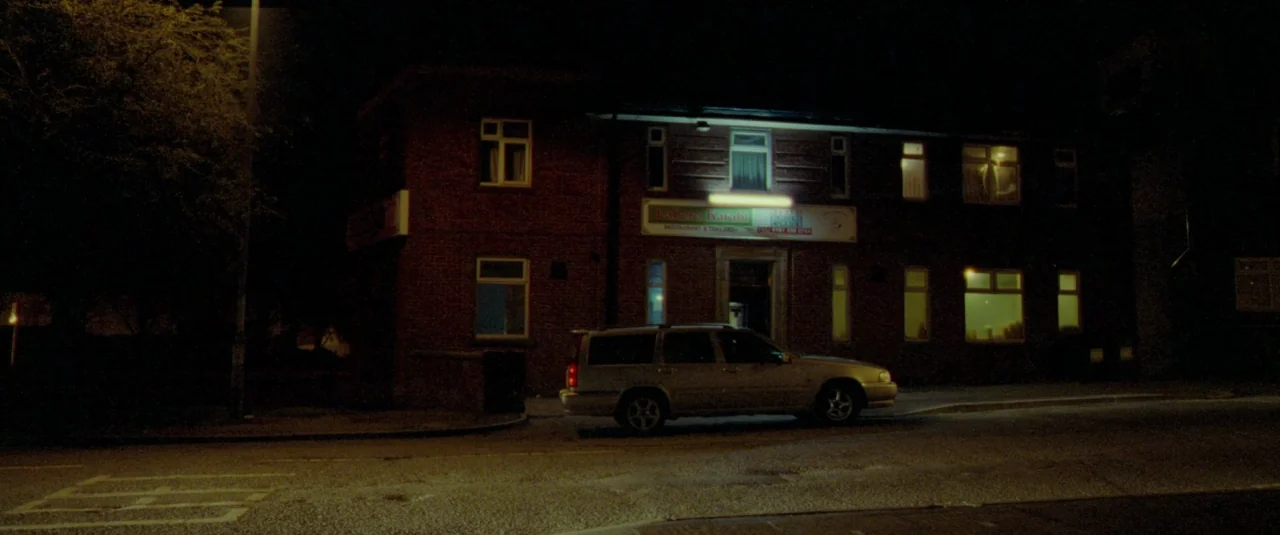 Catch Me Daddy (2014) movie still: establishing — A two-story red brick commercial building sits on a street corner at night. A silver station wagon…