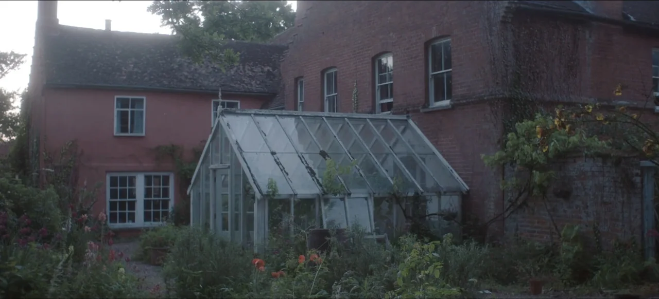 We Go In at Dawn (2020) movie still: wide — An old greenhouse sits nestled in a overgrown garden next to a brick house. The garden contains various…