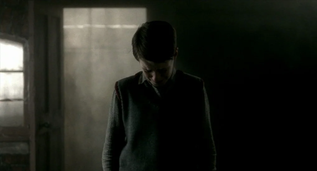 The Long Day Closes (1992) movie still: medium — A dimly lit, dusty corridor contains a boy standing in shadows. He faces downward with his head…