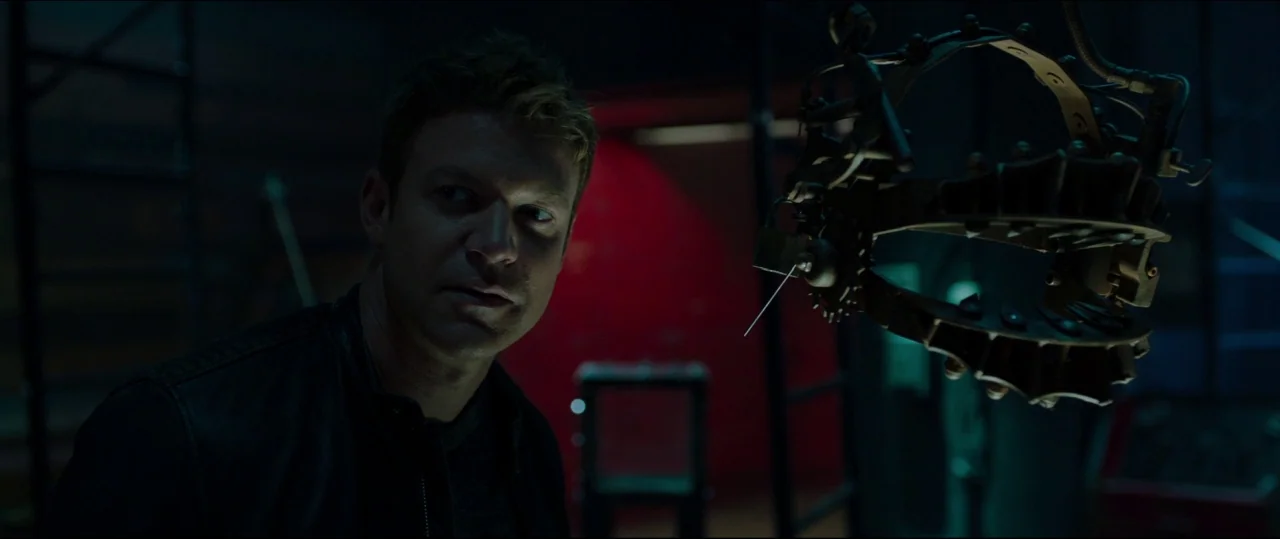 Jigsaw (2017) movie still: medium — A dark industrial basement area features dim blue shadows and a bright red background element. A man in a dark…
