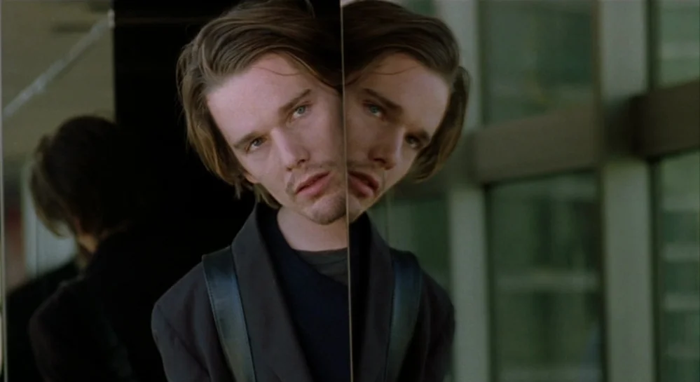 Hamlet (2000) movie still: closeup — A glass corridor interior features a vertical reflection splitting the frame. A young man gazes into the glass…